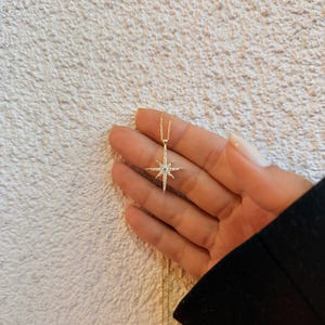 May include: A gold-colored necklace with a starburst pendant. The pendant is encrusted with small, clear gemstones. The necklace is held in a hand against a textured, off-white background. The starburst design has multiple points.