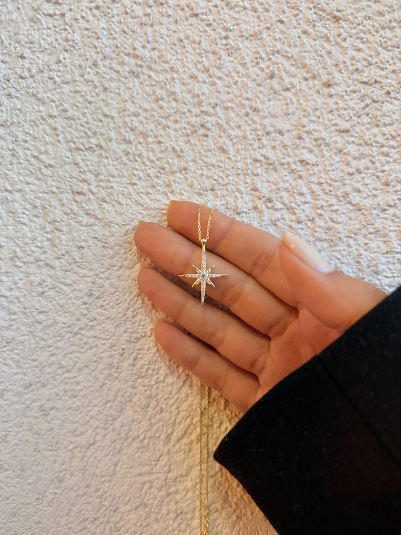 May include: A gold-colored necklace with a starburst pendant. The pendant is encrusted with small, clear gemstones. The necklace is held in a hand against a textured, off-white background. The starburst design has multiple points.