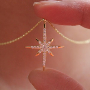 May include: A gold-colored necklace featuring a starburst pendant. The pendant is encrusted with small, clear gemstones and has pointed rays. The necklace is held between two fingers, showcasing the delicate chain.