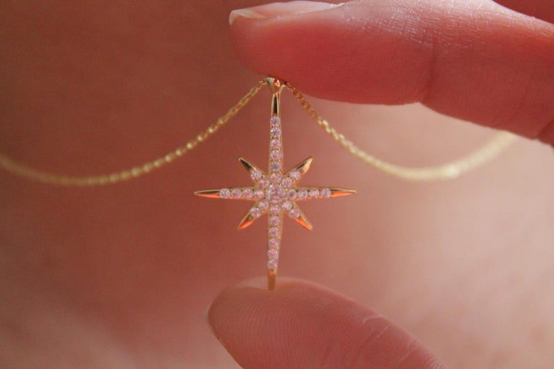 May include: A gold-colored necklace featuring a starburst pendant. The pendant is encrusted with small, clear gemstones and has pointed rays. The necklace is held between two fingers, showcasing the delicate chain.