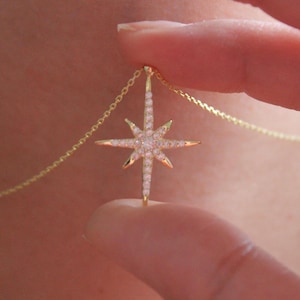 May include: A gold-colored necklace featuring a starburst pendant. The pendant is encrusted with small, sparkling crystals. The necklace is held by a person's fingers, showcasing the delicate chain and the pendant's intricate design. The starburst pendant is approximately 1 inch in diameter.
