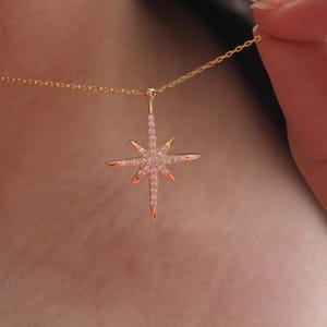 May include: A gold-colored necklace featuring a starburst pendant. The pendant is adorned with small, pink gemstones and has pointed edges. The necklace is being held up by a person's hand.