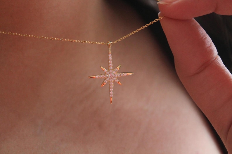 May include: A gold-colored necklace featuring a starburst pendant. The pendant is adorned with small, pink gemstones and has pointed edges. The necklace is being held up by a person's hand.