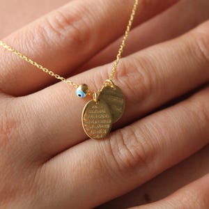 May include: A gold-tone chain necklace featuring two small oval-shaped pendants with engraved text and a tiny bead charm designed with a blue and white evil eye motif. The delicate metal chain connects to the charms, which hang together as a focal point.