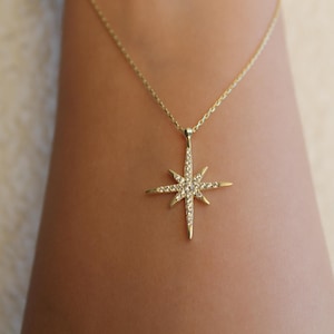 May include: A gold-colored necklace with a starburst pendant. The pendant is encrusted with small, sparkling gemstones. The necklace is displayed against a skin-toned background, highlighting the jewelry's design and color.