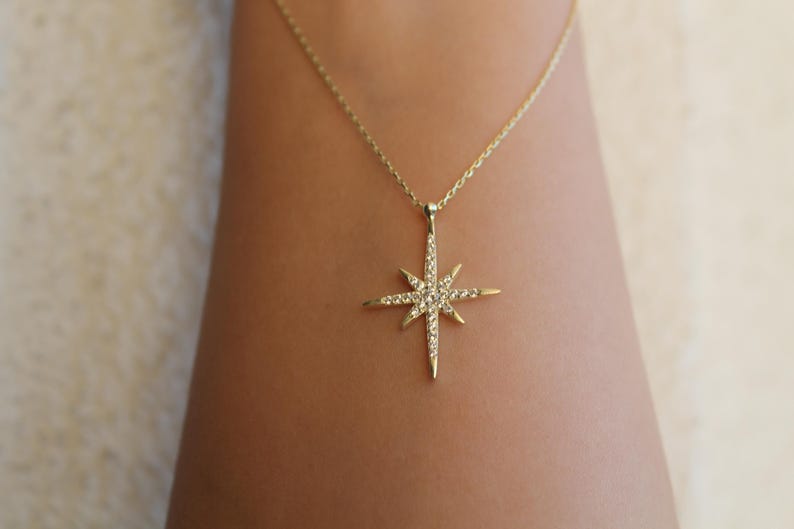 May include: A gold-colored necklace with a starburst pendant. The pendant is encrusted with small, sparkling gemstones. The necklace is displayed against a skin-toned background, highlighting the jewelry's design and color.