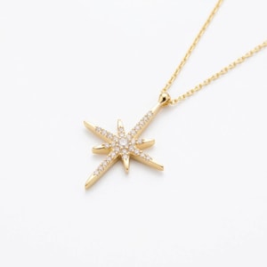 May include: A gold-colored necklace with a starburst pendant. The pendant is adorned with small, clear gemstones and is suspended from a delicate chain. The necklace is set against a white background.
