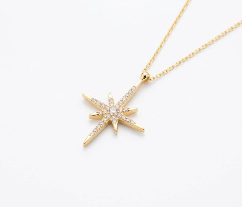 May include: A gold-colored necklace with a starburst pendant. The pendant is adorned with small, clear gemstones and is suspended from a delicate chain. The necklace is set against a white background.