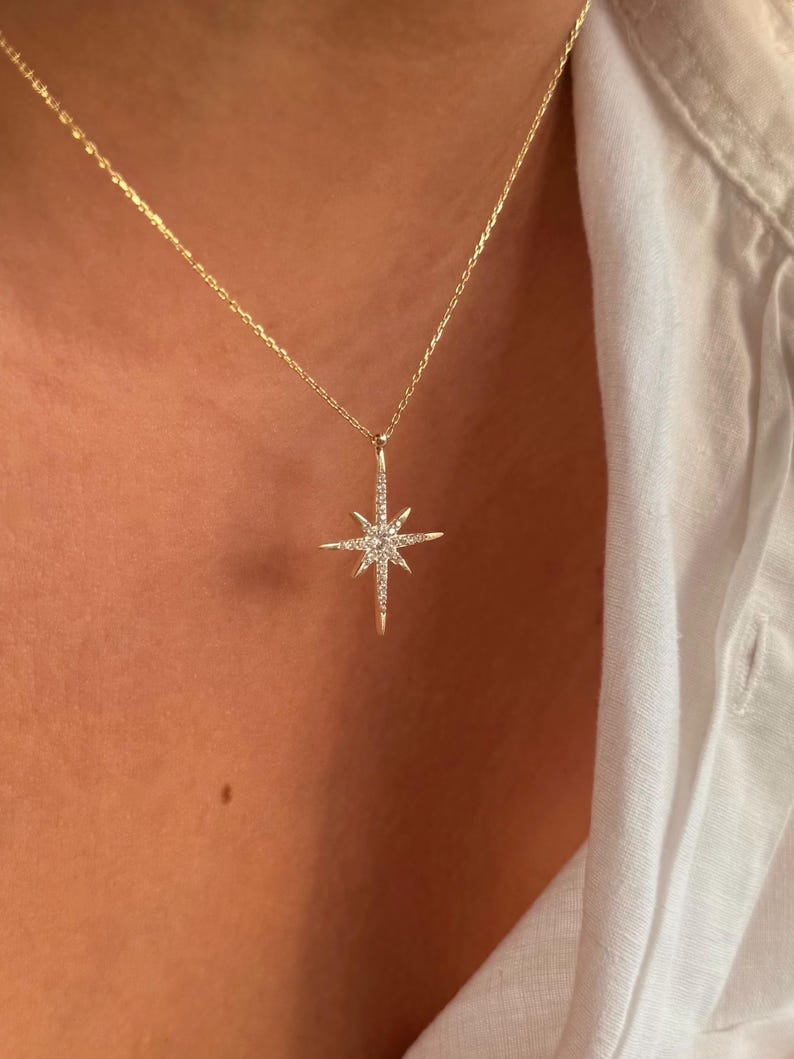 May include: A gold-colored necklace with a starburst pendant. The pendant is encrusted with small, sparkling gemstones. The necklace is worn against a person's skin and a white shirt.