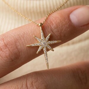 May include: A gold necklace featuring a starburst pendant with eight points of varying lengths. The pendant is encrusted with small, clear, sparkling stones across its surface and is suspended from a delicate, thin gold chain link necklace.