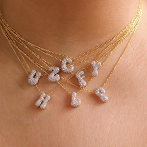 Könnte beinhalten: Ein Set aus geschichteten Goldketten, wobei jede feine Kette mit einem kleinen, abgerundeten, silberfarbenen Buchstabenanhänger versehen ist, der mit winzigen klaren Steinen besetzt ist. Die sichtbaren Buchstaben sind U, Z, G, F, Y, H, L und P.