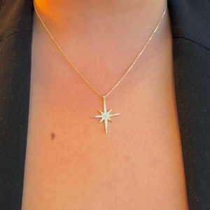 May include: A delicate gold necklace featuring a starburst pendant adorned with small, sparkling gemstones. The pendant hangs from a fine gold chain. The necklace is worn against a dark background.