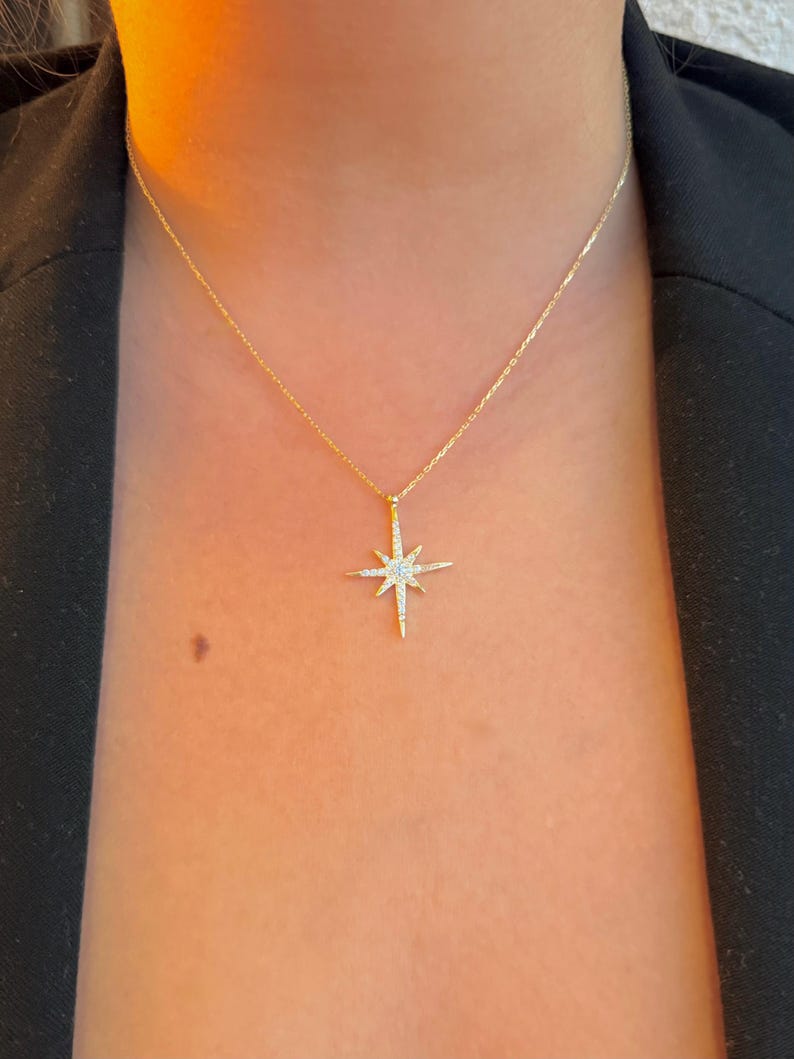 May include: A delicate gold necklace featuring a starburst pendant adorned with small, sparkling gemstones. The pendant hangs from a fine gold chain. The necklace is worn against a dark background.