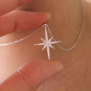 May include: A silver necklace featuring a starburst pendant. The pendant is encrusted with small, sparkling stones. The necklace is being held up, showcasing the design and detail of the jewelry.