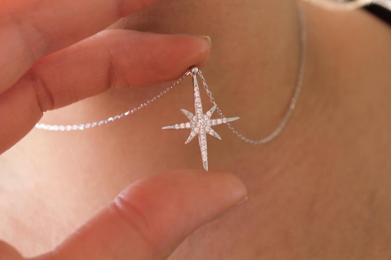 May include: A silver necklace featuring a starburst pendant. The pendant is encrusted with small, sparkling stones. The necklace is being held up, showcasing the design and detail of the jewelry.
