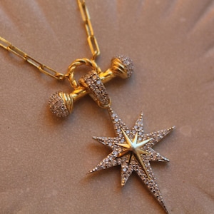 May include: A gold necklace with a starburst pendant, featuring a central gold element and small, sparkling stones. The necklace has a delicate chain and a distinctive clasp. The jewellery is displayed on a textured, neutral-toned surface.