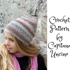 May include: A crochet hat with a striped pattern in shades of grey, pink, and white. The hat is worn by a person with long blonde hair. The text "Crochet Pattern by Capitanana Uncino" is visible in the image.