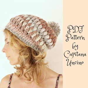 Può includere: Un cappello a beanie lavorato a uncinetto con un pon pon in tonalità beige, rosa e bianco. Il cappello è indossato da una persona con capelli biondi ricci. Il testo "PDF Pattern by Capitana Uncino" è visibile nell'immagine.