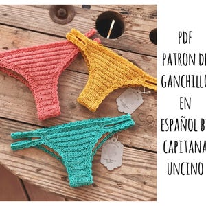 May include: Three crochet bikini bottoms in pink, yellow, and teal on a wooden surface. The image includes the text "PDF PATRON DE GANCHILLO EN ESPAÑOL BY CAPITANA UNCINO".