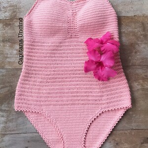 K&ouml;nnte beinhalten: Ein rosa geh&auml;kelter Badeanzug mit einem gewellten Ausschnitt und einer Blumenverzierung. Der Badeanzug ist aus feinem Garn gefertigt und hat einen zarten, femininen Look.