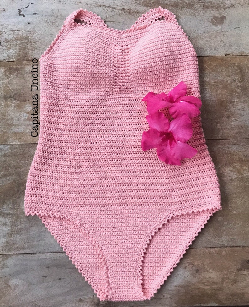 K&ouml;nnte beinhalten: Ein rosa geh&auml;kelter Badeanzug mit einem gewellten Ausschnitt und einer Blumenverzierung. Der Badeanzug ist aus feinem Garn gefertigt und hat einen zarten, femininen Look.