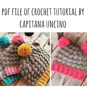 May include: Two crocheted hats with colorful pom-poms. The hats are gray with a pink and yellow band at the bottom. The hats are on a wooden surface. PDF FILE OF CROCHET TUTORIAL BY CAPITANA UNCINO.