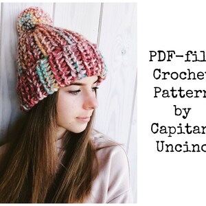 Può includere: Un cappello all'uncinetto colorato con un pon pon in cima. Il cappello è realizzato con una varietà di colori, tra cui rosa, blu e giallo. Il cappello è indossato da una persona con i capelli lunghi e castani. Il testo "PDF-file Crochet Pattern by Capitanana Uncino" è visibile nell'immagine.
