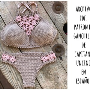 May include: A crochet bikini top and bottom in beige with pink floral accents. The pattern is called "Capitana Uncino" and is written in Spanish.