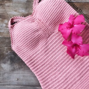K&ouml;nnte beinhalten: Ein rosa geh&auml;keltes Oberteil mit einem gewellten Rand. Das Oberteil liegt auf einer Holzoberfl&auml;che mit rosa Bl&uuml;tenbl&auml;ttern.