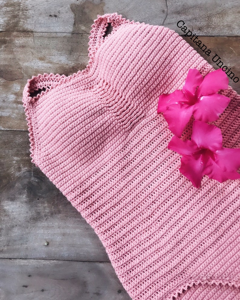 K&ouml;nnte beinhalten: Ein rosa geh&auml;keltes Oberteil mit einem gewellten Rand. Das Oberteil liegt auf einer Holzoberfl&auml;che mit rosa Bl&uuml;tenbl&auml;ttern.