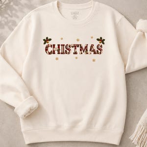 May include: A cream-colored crewneck sweatshirt featuring the text CHISTMAS in a red and white plaid pattern. The design is accented with small gold snowflakes and two sprigs of holly with red berries positioned above the letters.