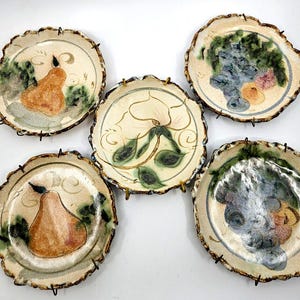 May include: Five decorative ceramic plates with organic, irregular edges. Each plate features a unique hand-painted design of fruit or floral motifs in shades of orange, green, blue, and pink. The plates are mounted on black metal stands.