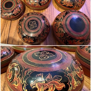 May include: A collection of decorative, round boxes with intricate designs. The boxes feature a black base with red and gold floral patterns. The lids have a central black circle. The boxes are arranged on a wooden surface.