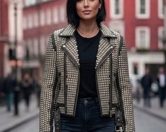 May include: A studded, olive green and silver biker jacket with a notched collar, asymmetrical zipper, and belt. The jacket is worn over a black top and dark wash jeans. The background shows a city street with red buildings.
