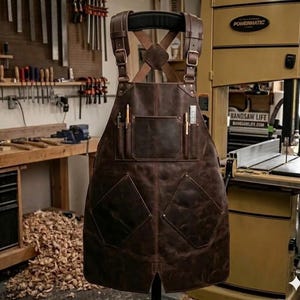 Puede incluir: Delantal de cuero marrón con múltiples bolsillos y correas ajustables. El delantal tiene un diseño cruzado en la espalda y se muestra en un taller. Cuenta con bolsillos para herramientas y un portalápices. Diseñado para carpintería.
