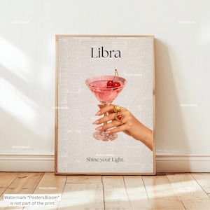 May include: A framed print featuring a pink cocktail with cherries, held by a hand adorned with rings. The word "Libra" is at the top, with "Shine your Light" below. The background has a newspaper print design.