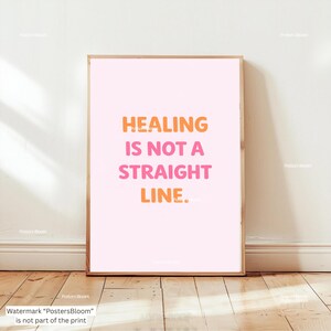 Könnte beinhalten: Ein gerahmter Druck mit dem Text "HEALING IS NOT A STRAIGHT LINE" in Orange und Pink auf einem hellrosa Hintergrund. Der Druck befindet sich in einem Holzrahmen und wird auf einem Holzboden präsentiert.