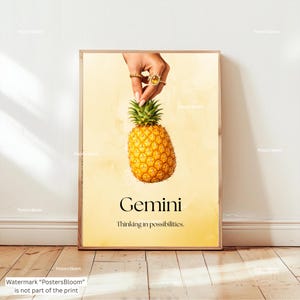 May include: A framed print featuring a pineapple held by a hand with rings, set against a yellow background. The word "Gemini" is displayed in black text, with the phrase "Thinking in possibilities." below it. The print has a wooden frame.