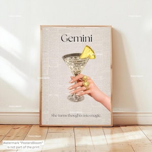 May include: A framed print with a cocktail in a martini glass and pineapple garnish. The word "Gemini" is at the top, with the phrase "She turns thoughts into magic" below. A hand wearing gold rings holds the glass.
