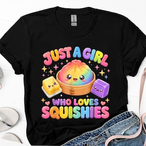 Może przedstawiać: Czarny t-shirt z krótkim rękawem z kolorową grafiką przedstawiającą tęczowego pierożka w bambusowym koszyku do gotowania na parze, żółtą kostkę masła i fioletową kostkę. Napis głosi JUST A GIRL WHO LOVES SQUISHIES zaokrągloną czcionką.