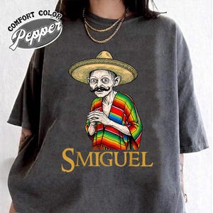 May include: A charcoal grey t-shirt featuring a graphic of a character wearing a tan sombrero, a black moustache, and a colourful striped poncho. The text "SMIGUEL" is printed in gold below the graphic, and a "COMFORT COLOR Pepper" logo is at the top.