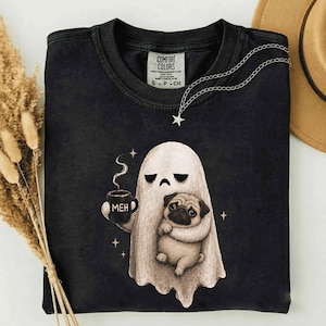 May include: A black crewneck t-shirt featuring a graphic of a white ghost holding a black mug with the text MEH printed on it. The ghost is embracing a tan pug. The shirt has a Comfort Colors brand label on the inside of the collar area.