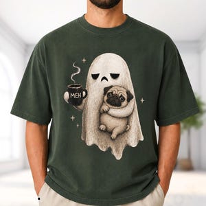 Puede incluir: Una camiseta de color verde bosque oscuro con una ilustración gráfica de un fantasma blanco que sostiene una taza negra con el texto MEH impreso. El fantasma abraza a un perro pug beige y hay pequeñas estrellas de cuatro puntas alrededor.