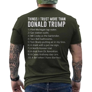Op de afbeelding: Een olijfgroen t-shirt met witte tekst op de achterkant die luidt: THINGS I TRUST MORE THAN DONALD TRUMP, gevolgd door een genummerde lijst van tien humoristische items, waaronder kraanwater uit Flint Michigan, tankstationsushi en een scheet.