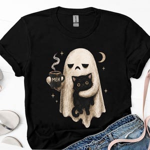 May include: A black short-sleeved t-shirt featuring a graphic of a beige ghost holding a black mug with the text MEH and a black cat. The ghost has a bored expression and is surrounded by small stars and a crescent moon on the chest area.