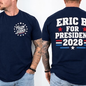 May include: A navy blue short-sleeve t-shirt featuring a front chest graphic with the text Follow The Leader surrounded by stars. The back displays the text ERIC B. FOR PRESIDENT 2028 in white with red stars and red and blue horizontal stripes.