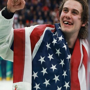 May include: A person smiling and celebrating, draped in an American flag. The flag has red and white stripes and white stars on a blue field. The person is wearing a white jacket.