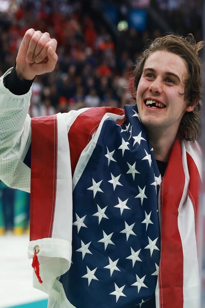 May include: A person smiling and celebrating, draped in an American flag. The flag has red and white stripes and white stars on a blue field. The person is wearing a white jacket.