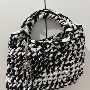 May include: A handmade handbag crafted from thick black and white fabric yarn in a chunky crochet stitch. The bag features a single integrated handle and a silver-toned metal charm hanging from the side, depicting a dreamcatcher with small colorful beads.
