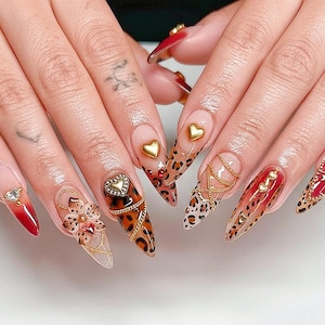 Puede incluir: Un conjunto de uñas largas y puntiagudas con varios diseños. Algunas uñas presentan un estampado de leopardo, cadenas doradas y adornos en forma de corazón. Otras tienen un diseño degradado rojo y transparente con detalles de pedrería. Las uñas están hechas profesionalmente.